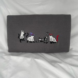 May include: A gray sweatshirt with an embroidered design of five cats dressed as ghosts and a skeleton. The cats are in a line, with the skeleton cat in the middle. The ghosts are wearing hats, and the skeleton cat has a red devil tail.