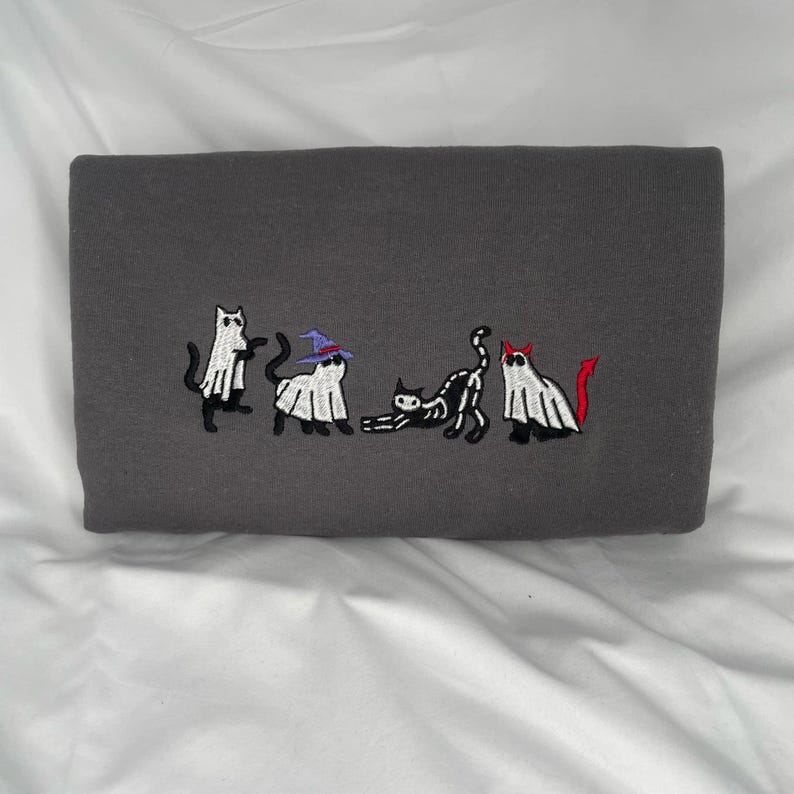 May include: A gray sweatshirt with an embroidered design of five cats dressed as ghosts and a skeleton. The cats are in a line, with the skeleton cat in the middle. The ghosts are wearing hats, and the skeleton cat has a red devil tail.
