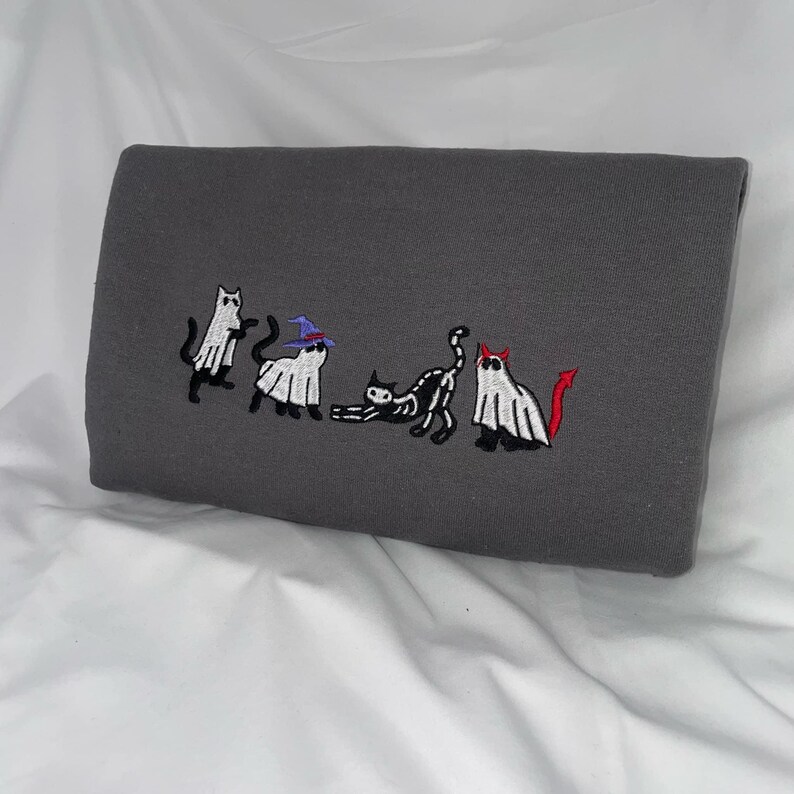 May include: A gray sweatshirt with an embroidered design of five cats dressed as ghosts for Halloween. One cat wears a witch's hat, another has a devil's tail, and the others are plain ghosts.