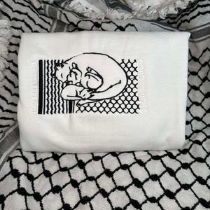 May include: White embroidered t-shirt featuring a black line drawing of two cats cuddling within a black and white geometric pattern.