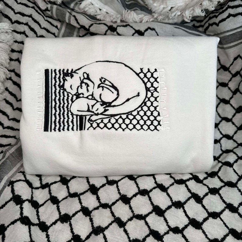 May include: White embroidered t-shirt featuring a black line drawing of two cats cuddling within a black and white geometric pattern.