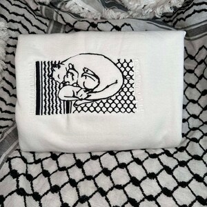 May include: White t-shirt featuring a black embroidered design of two cats cuddling. The design is framed by a black and white geometric pattern.