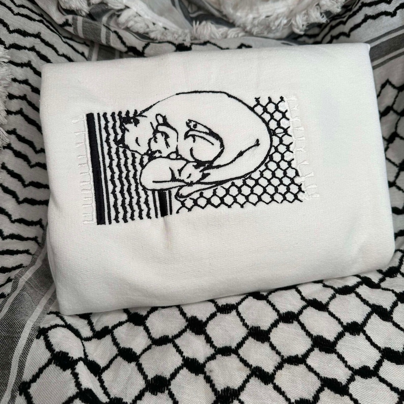 May include: White t-shirt featuring a black embroidered design of a cat curled up with a smaller cat. The design is enclosed within a black and white grid pattern.