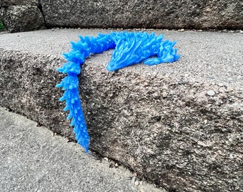 Puede incluir: Una figura de dragón azul, impresa en 3D, con espinas y una cola larga. El dragón está acostado en un conjunto de escalones de concreto.