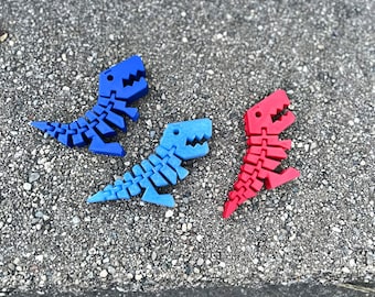 Puede incluir: Tres esqueletos de dinosaurio de plástico, uno azul, uno rojo y uno azul claro, están dispuestos sobre una superficie gris y de guijarros.