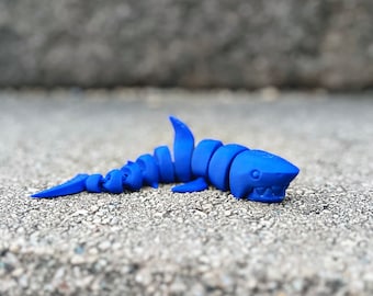 Puede incluir: Un tiburón de juguete de plástico azul con un cuerpo segmentado. El tiburón está acostado sobre una superficie de concreto gris.