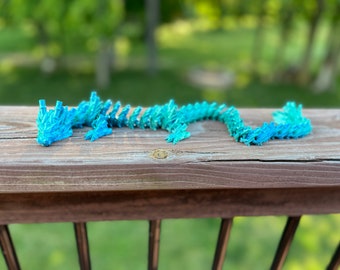 Puede incluir: Un dragón de origami azul y verde hecho de papel. El dragón está colocado sobre una superficie de madera.