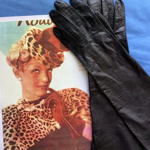 Può includere: Un paio di guanti lunghi in pelle nera con una finitura liscia e leggermente testurizzata. I guanti presentano un design classico che si estende oltre il polso, mostrando cuciture visibili lungo le dita e un aspetto morbido e flessibile.