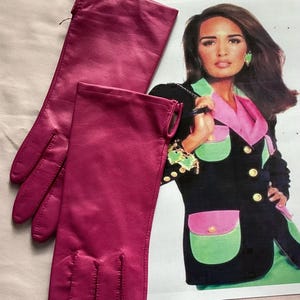 May include: A pair of long, solid magenta gloves with visible stitching along the fingers and a smooth finish. The gloves are positioned next to a printed image of a person wearing a black blazer with pink and green pocket details.