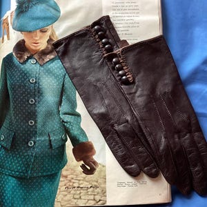 May include: A pair of dark brown leather gloves featuring a row of five small, round, matching buttons along the outer wrist area. The gloves have visible stitched detailing on the back of the hand and a smooth, matte finish on the leather surface.