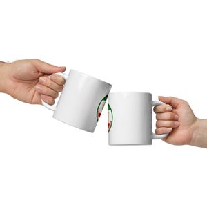 Może przedstawiać: Dwa białe ceramiczne kubki z uchwytem są trzymane w geście toastu. Każdy kubek ma okrągły wzór z czerwonymi, zielonymi i złotymi akcentami. Kubki są na białym tle.