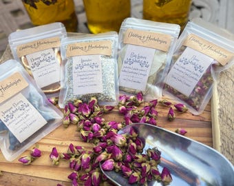 Op de afbeelding: Een assortiment gedroogde kruiden en botanische producten, waaronder rozenknoppen en lavendel, wordt gepresenteerd in doorzichtige zakjes en glazen potten. Een metalen schep bevat een stapel roze rozenknoppen. De tekst "Oddities & Herbologies" is zichtbaar.