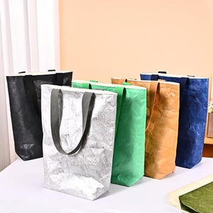 以下が含まれることがあります： 黒、シルバー、緑、タン、青など、さまざまな色合いのカラフルなトートバッグのコレクション。各バッグは長方形で、お揃いのハンドルが付いています。バッグは質感のある素材で作られています。