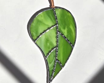 May include: A stained glass leaf ornament in shades of green, with a silver-colored metal border and internal veins. A brown leather cord is attached for hanging. The leaf design is reminiscent of nature.