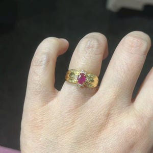 Puede incluir: Un anillo de oro con una gran piedra central de rubí ovalada, rodeada de pequeñas piedras blancas. El anillo tiene una banda detallada y texturizada con piedras pequeñas adicionales. El anillo está en un dedo.