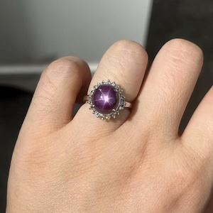 May include: A silver ring featuring a large, round, purplish-red gemstone with a six-pointed star pattern. The gemstone is surrounded by a halo of smaller, clear stones. The ring is on a hand.