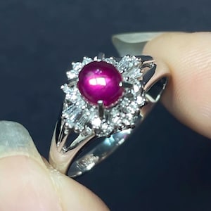 May include: A silver-toned ring featuring a central oval cabochon-cut deep pink gemstone. The stone is surrounded by a cluster of small, clear, faceted white stones arranged in a halo setting with additional baguette-cut stones on the sides.