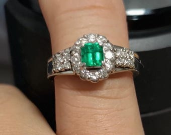 Op de afbeelding: Een zilveren ring met een vierkant geslepen, levendig groene smaragd als middensteen, omringd door een halo van kleinere, heldere diamanten. Extra diamanten zijn gezet op de band van de ring, wat een sprankelend, elegant ontwerp creëert.
