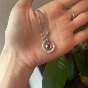 May include: Silver pendant necklace featuring a teardrop-shaped design. The pendant has a central oval-shaped red gemstone, surrounded by small, clear stones. The necklace chain is delicate and silver-colored.