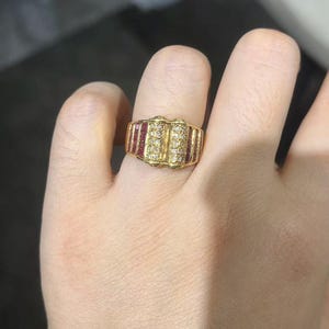 May include: A gold ring with a rectangular design. The ring features a central section of small, clear stones, flanked by rows of red stones. The ring is set on a finger.