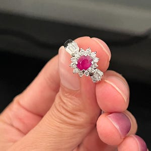 May include: A silver-toned ring featuring a central oval-cut vibrant red ruby gemstone surrounded by a halo of small round white diamonds. The band is accented with baguette-cut diamonds on each side of the central cluster setting.