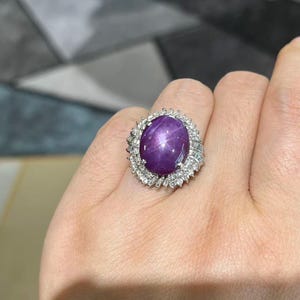 May include: A ring featuring a large, oval-shaped, deep purple star gemstone at the center. The stone displays a distinct six-rayed star pattern. It is surrounded by a halo of small, clear, sparkling baguette and round-cut white stones.