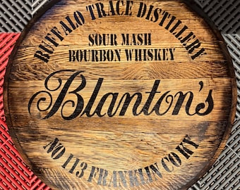 May include: Round wooden sign with the text "Buffalo Trace Distillery" and "Blanton's" in black lettering. The sign also includes "Sour Mash Bourbon Whiskey" and "No 113 Franklin Co KY". The wood has a natural grain.