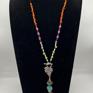 May include: A long necklace featuring a strand of orange, pink, white, and lime green beads. A silver-toned metal leaf charm hangs from a toggle clasp, followed by a patterned bead, a light blue skull-shaped bead, a red rose, and a gold triangle.