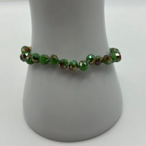 May include: A beaded bracelet featuring faceted, round green glass beads with an iridescent finish. The beads are strung together in a single strand, displaying a reflective surface that shifts between shades of bright green and metallic bronze tones.