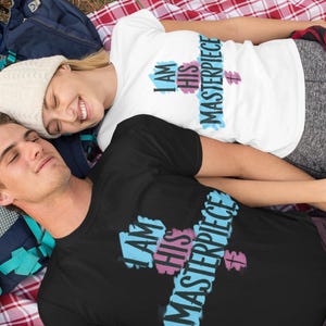 May include: A couple wearing matching black and white t-shirts with the text "I am his masterpiece" and "I am his masterpiece" on them. The couple is lying on a red and white checkered blanket.