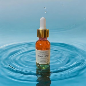 May include: A clear glass bottle with a gold cap and dropper filled with a light orange liquid. The bottle is floating in a blue water ripple and has a label that reads "Everlasting Youth Serum" and "Handcrafted by Skin & Beyond Essentials".