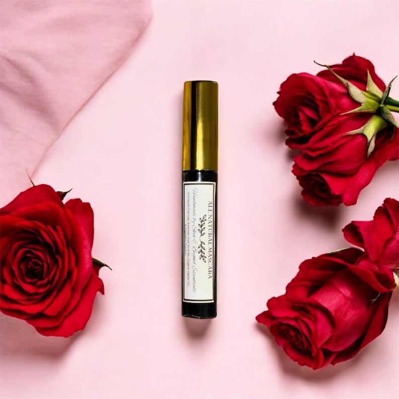May include: A black mascara tube with a gold cap and a white label that reads "All Natural Mascara" with a small leaf design. The tube is surrounded by three red roses on a pink background.
