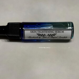 Pu&ograve; includere: Un piccolo flacone di vetro blu con un contagocce e un'etichetta bianca che recita "SKIN TIGHTENING SERUM Handmade by Skin & Beyond Essentials".