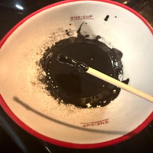 May include: A white ceramic bowl with a red rim and the words "ONE CUP" printed on the bottom. The bowl contains a black liquid and a wooden stick.