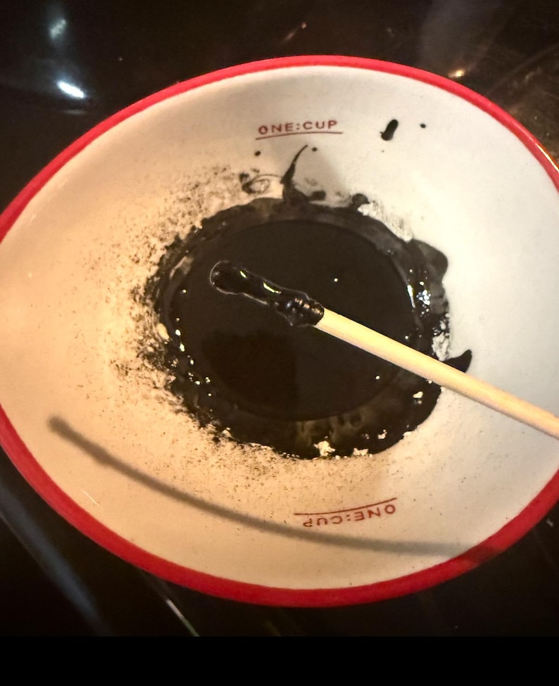May include: A white ceramic bowl with a red rim and the words "ONE CUP" printed on the bottom. The bowl contains a black liquid and a wooden stick.