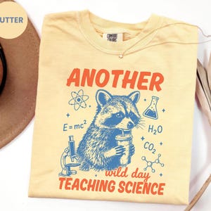 May include: A butter yellow t-shirt featuring a graphic of a raccoon holding a drink, surrounded by science-themed icons including a microscope, an atom, a beaker, and chemical formulas, with the text "ANOTHER WILD DAY TEACHING SCIENCE".