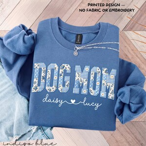 May include: An indigo blue crewneck sweatshirt featuring the text DOG MOM in a blue and white gingham and floral pattern. Below, the names daisy and lucy are written in white script with a heart. Text reads PRINTED DESIGN — NO FABRIC OR EMBROIDERY.