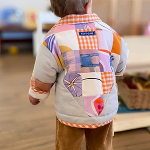 May include: A child wearing a light blue quilted jacket with a patchwork design featuring various colors and patterns, including orange, pink, purple, and yellow. The jacket has a label that reads "PATTI-CAKE". The child is also wearing brown corduroy pants.