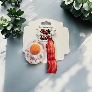 May include: A pair of mismatched dangle earrings featuring a fried egg with a bright orange yolk and a strip of crispy bacon. The earrings are attached to a white display card printed with the text Bizzy Beez Art Studio and a bee logo.