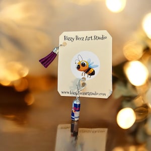 May include: A miniature Red Bull can charm with a purple tassel and a tag featuring a cartoon bee and the text "Bizzy Beez Art Studio" and "www.bizzybeezartstudio.com". The charm is attached to a silver-colored clasp.