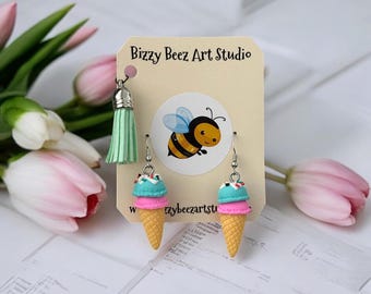 May include: A pair of dangle earrings shaped like ice cream cones with teal and pink scoops on a Bizzy Beez Art Studio display card. The card features a cartoon bee illustration and a light green tassel charm attached to the top left corner.