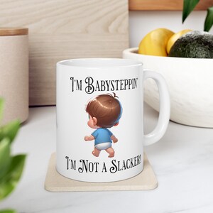May include: White ceramic mug with a black and white illustration of a baby taking its first steps. The text on the mug reads "I'm babysteppin' I'm not a slacker!"