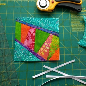 May include: A colorful patchwork quilt square with vibrant green, orange, pink, and teal fabrics. The square is on a green cutting mat with a rotary cutter, ruler, and white strips of fabric. The quilt block features angled stripes.