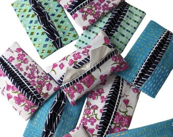 May include: A selection of fabric tissue box covers in diverse patterns. Some covers display floral designs with pink flowers on a white backdrop, while others have geometric or dotted patterns in shades of blue and green. Each cover has a contrasting trim.