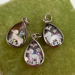 May include: Three silver-toned teardrop pendants with a ram design. Each pendant features a white ram with a detailed design inside a clear, iridescent background. The pendants are displayed on a green velvet surface.
