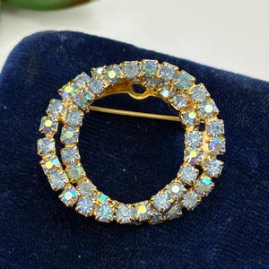 May include: A vintage gold-tone brooch with a double-circle design. The circles are encrusted with light blue, iridescent rhinestones. The pin has a classic, elegant style, perfect for adding a touch of sparkle to any outfit.