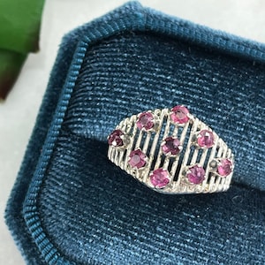 May include: A silver ring with a geometric design featuring pink gemstones. The ring is set in a blue velvet ring box.