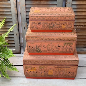 May include: Three stacked wooden boxes with intricate hand-painted designs. The boxes are decorated with scenes of people and animals in a traditional style. The boxes are painted in shades of pink, orange, and green.