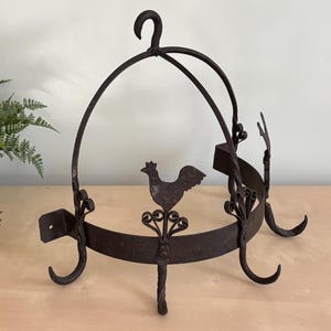 May include: A black metal pot rack with a rooster design and multiple hooks. The rack is designed to hang from the ceiling.
