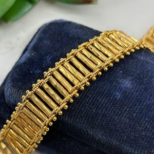 May include: Close-up of a gold-colored bracelet with a textured, rectangular design. The bracelet is resting on a dark blue velvet surface. The bracelet's design features a series of parallel, textured bars. The edges are adorned with small, round beads.